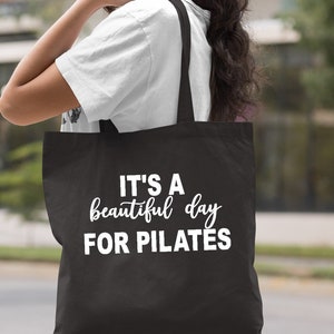 May include: Black tote bag with the text "IT'S A beautiful day FOR PILATES" in white. The bag has a simple design and is carried over the shoulder. The word "Black" is printed at the bottom right.