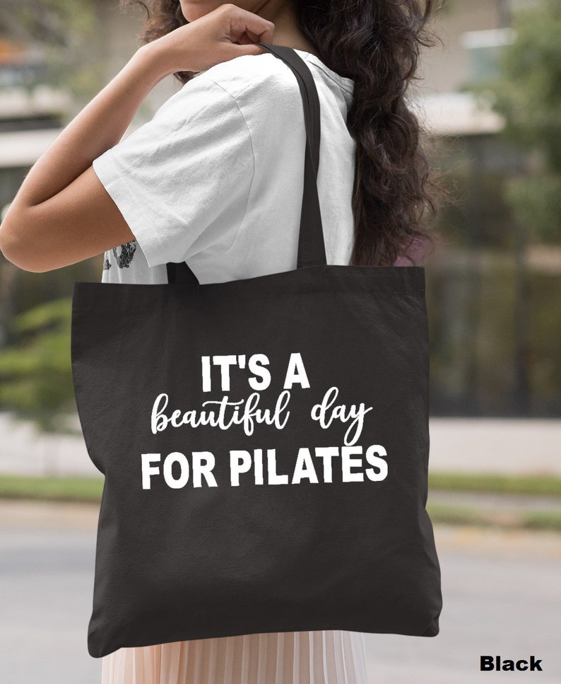 May include: Black tote bag with the text "IT'S A beautiful day FOR PILATES" in white. The bag has a simple design and is carried over the shoulder. The word "Black" is printed at the bottom right.