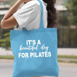 May include: Light blue tote bag with white text that reads "IT'S A beautiful day FOR PILATES". The bag is carried over the shoulder. The background is blurred, suggesting an outdoor setting.