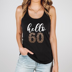 May include: Black tank top with the words "hello 60" in white script. The number 60 is in a leopard print pattern. The model is wearing blue jeans and a gold necklace.