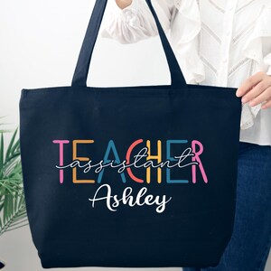 May include: A navy blue tote bag with black handles. The bag features the words "TEACHER assistant Ashley" in colorful, stylized lettering. The bag is held by a person wearing a white blouse and blue jeans.