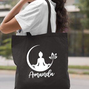 May include: Black canvas tote bag with a white silhouette of a person meditating in a crescent moon with a leaf design. The name "Amanda" is printed below the design.