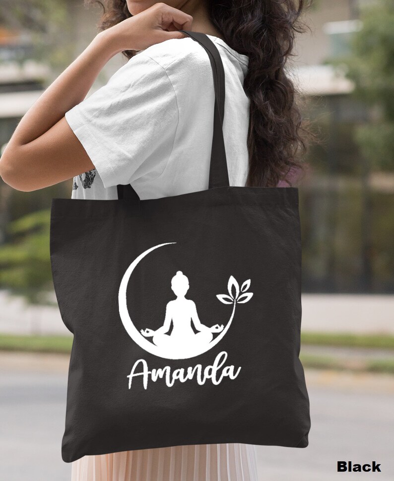 May include: Black canvas tote bag with a white silhouette of a person meditating in a crescent moon with a leaf design. The name "Amanda" is printed below the design.
