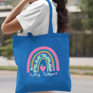 May include: A royal blue tote bag with a rainbow design featuring a heart, pencils, and an apple. The text "Mrs. Wilson" is printed on the bag.
