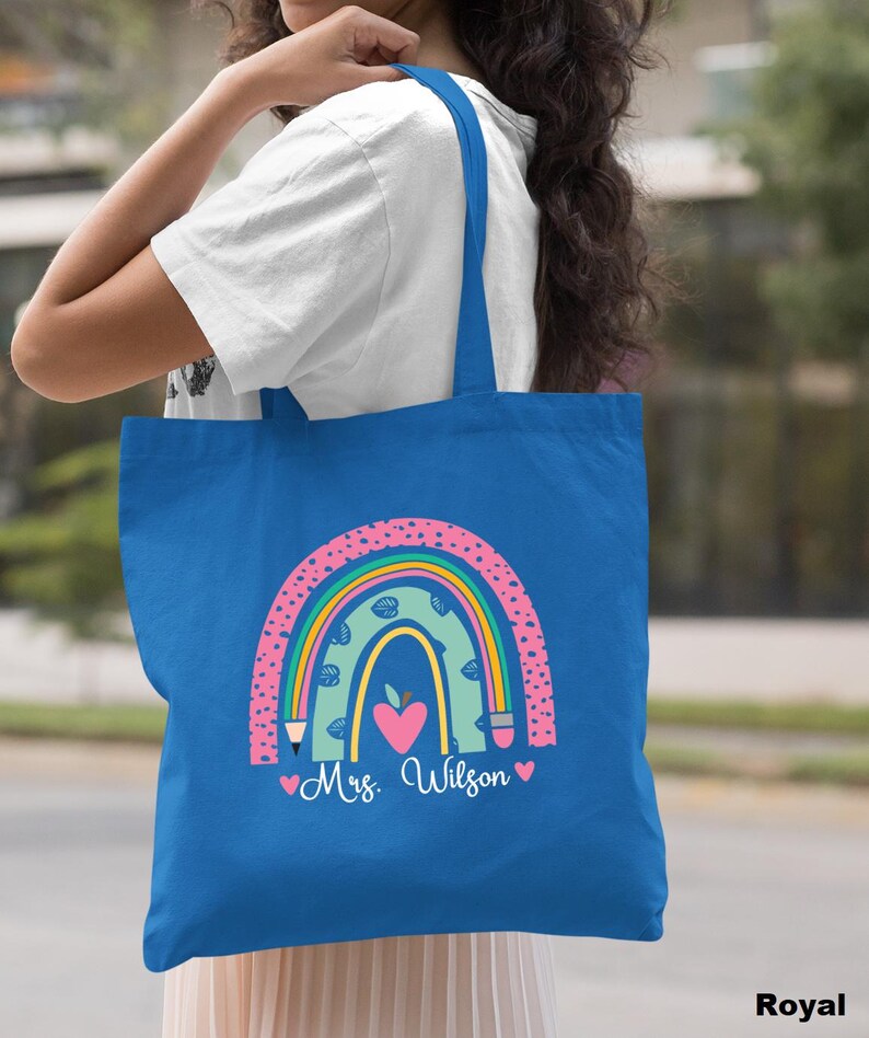 May include: A royal blue tote bag with a rainbow design featuring a heart, pencils, and an apple. The text "Mrs. Wilson" is printed on the bag.