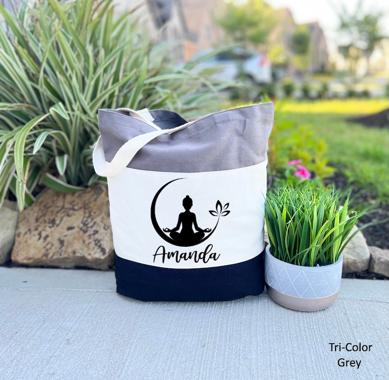 May include: A tri-color tote bag with a grey, white, and black design. The bag features a black silhouette of a person in a yoga pose with a lotus flower. The name "Amanda" is printed in black below the image.