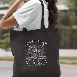 Peut inclure: Sac fourre-tout noir avec l'inscription blanche "HOMESCHOOL MAMA RAISING ARROWS". Le motif comprend un livre ouvert avec des illustrations florales et de papillons. Le sac est port&eacute; sur l'&eacute;paule.