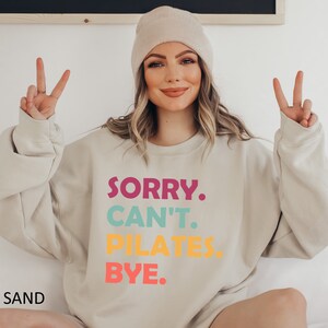 May include: A beige sweatshirt with the text "SORRY. CAN'T. PILATES. BYE." in colorful letters. The text is in a stacked format with each word in a different color. The sweatshirt is being worn by a person with a beige beanie.