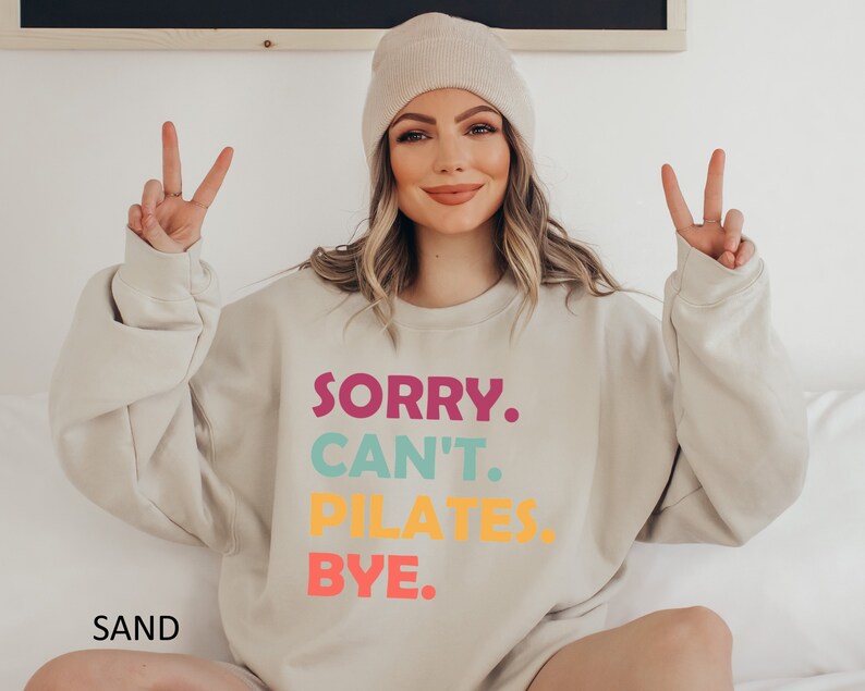 May include: A beige sweatshirt with the text "SORRY. CAN'T. PILATES. BYE." in colorful letters. The text is in a stacked format with each word in a different color. The sweatshirt is being worn by a person with a beige beanie.
