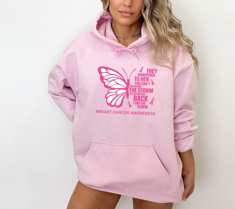 May include: Pink hoodie with a butterfly graphic and the text "THEY WHISPERED TO HER YOU CAN'T WITHSTAND THE STORM SHE WHISPERED BACK I AM THE STORM" and "BREAST CANCER AWARENESS".