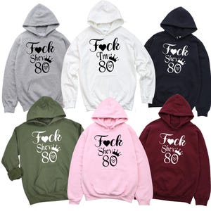 May include: Six different color hooded sweatshirts with the text "Fock She's 80" printed on the front. The sweatshirts are in grey, white, black, olive green, pink, and burgundy.