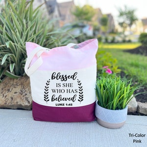 May include: A tri-color tote bag with a pink, white, and burgundy design. The bag has a white canvas panel with the text "blessed is she who has believed" and "LUKE 1:45" printed in black.