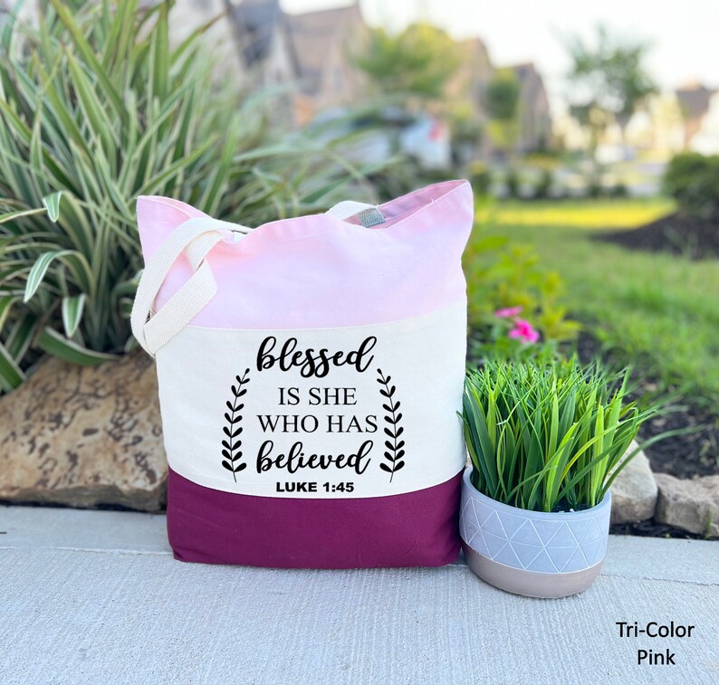 May include: A tri-color tote bag with a pink, white, and burgundy design. The bag has a white canvas panel with the text "blessed is she who has believed" and "LUKE 1:45" printed in black.