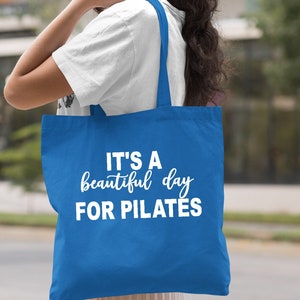 May include: Royal blue tote bag with white text that reads "IT'S A beautiful day FOR PILATES". The bag has two straps and is being held over a person's shoulder. The background is blurred.