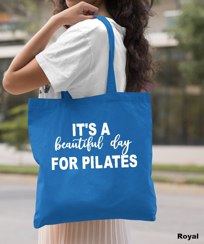 May include: Royal blue tote bag with white text that reads "IT'S A beautiful day FOR PILATES". The bag has two straps and is being held over a person's shoulder. The background is blurred.