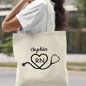 May include: A natural canvas tote bag with a black design of a stethoscope and a heart with the letters "RN" inside. The text "Sophia" is printed above the heart.