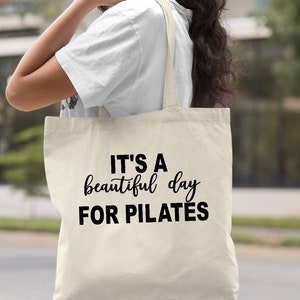 May include: A natural canvas tote bag with the text "IT'S A beautiful day FOR PILATES" in black script and block letters. The bag has long straps and is being held by a person. The word "Natural" is printed in the bottom right corner.