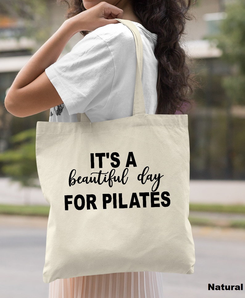 May include: A natural canvas tote bag with the text "IT'S A beautiful day FOR PILATES" in black script and block letters. The bag has long straps and is being held by a person. The word "Natural" is printed in the bottom right corner.