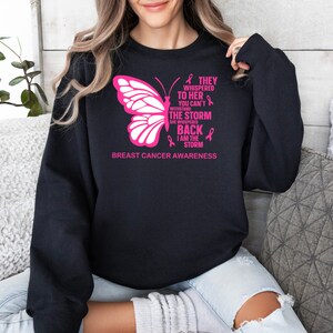 May include: Black sweatshirt with a pink butterfly and the text "They whispered to her you can't withstand the storm she whispered back I am the storm" and "Breast Cancer Awareness".