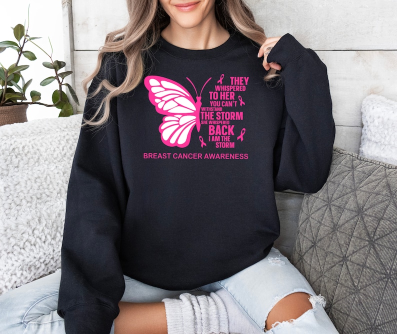 May include: Black sweatshirt with a pink butterfly and the text "They whispered to her you can't withstand the storm she whispered back I am the storm" and "Breast Cancer Awareness".