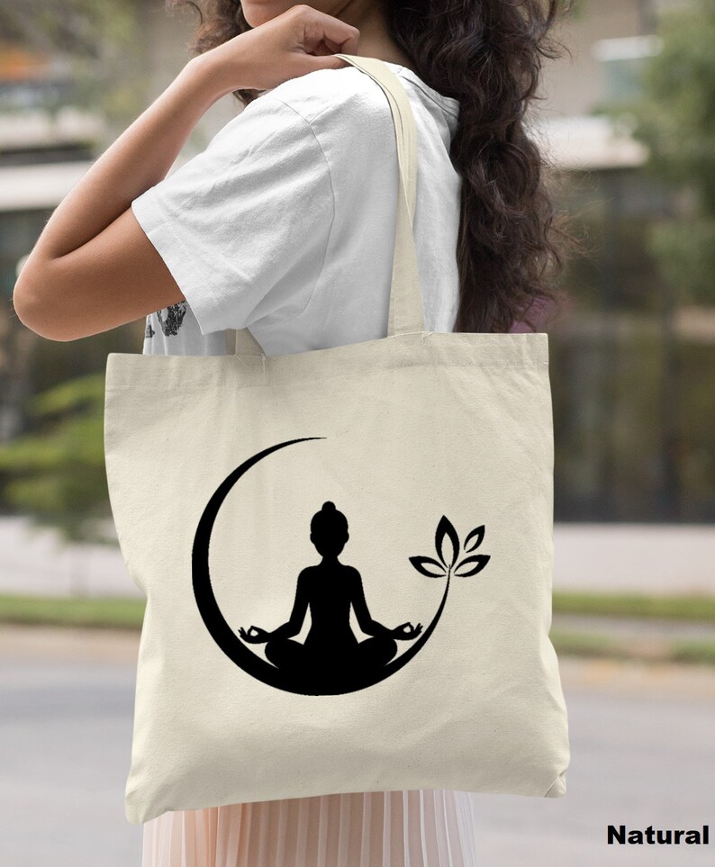May include: A natural canvas tote bag with a black silhouette of a person meditating in a crescent moon with a lotus flower. The text "Natural" is printed on the bottom right corner of the image.