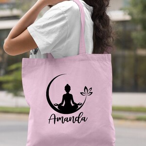 May include: Light pink tote bag with a black silhouette of a person meditating in a crescent moon with a lotus flower. The bag is personalized with the name "Amanda".