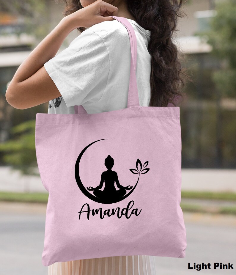 May include: Light pink tote bag with a black silhouette of a person meditating in a crescent moon with a lotus flower. The bag is personalized with the name "Amanda".