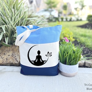 May include: A blue, white, and navy blue tote bag with a silhouette of a person meditating on a crescent moon with a lotus flower. The bag has a white canvas strap.