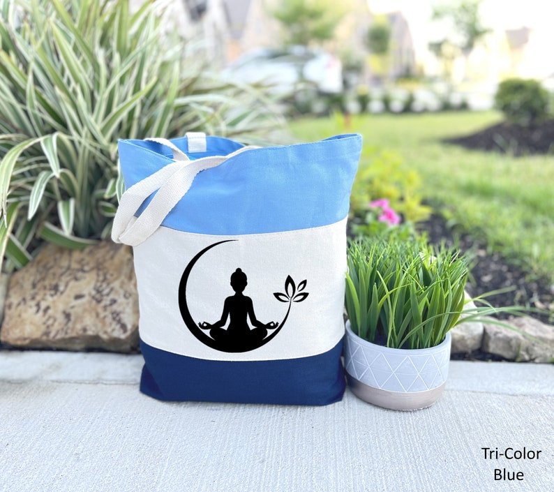 May include: A blue, white, and navy blue tote bag with a silhouette of a person meditating on a crescent moon with a lotus flower. The bag has a white canvas strap.