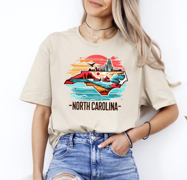 May include: A beige t-shirt with a graphic of the state of North Carolina. The graphic is a colorful illustration of the state's outline with a sunset, mountains, and a cityscape. The text "North Carolina" is printed below the graphic.