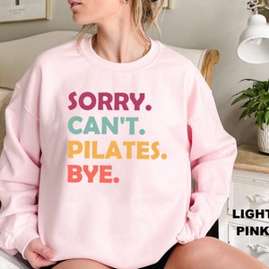 May include: A light pink sweatshirt with the text "SORRY. CAN'T. PILATES. BYE." in colorful letters.
