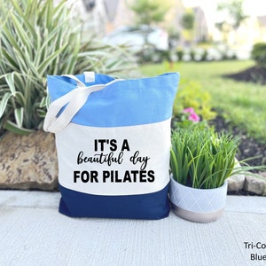 May include: A tri-color blue tote bag with white handles. The bag is divided into three horizontal sections: light blue, white, and navy blue. The text "IT'S A beautiful day FOR PILATES" is printed in black on the white section. The bag is designed for carrying items.