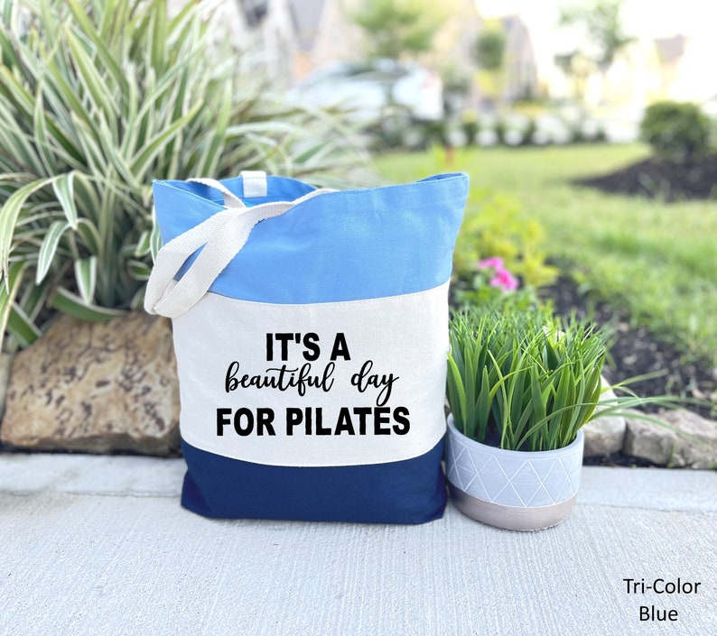 May include: A tri-color blue tote bag with white handles. The bag is divided into three horizontal sections: light blue, white, and navy blue. The text "IT'S A beautiful day FOR PILATES" is printed in black on the white section. The bag is designed for carrying items.