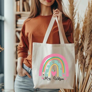 May include: A white canvas tote bag with a rainbow design and the text "Mrs. Wilson". The rainbow is made up of pink, green, yellow, and blue stripes with white polka dots. There is a red apple and a pencil on the rainbow.