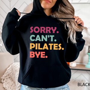 May include: Black hooded sweatshirt with the text "SORRY. CAN'T. PILATES. BYE." in colorful letters.