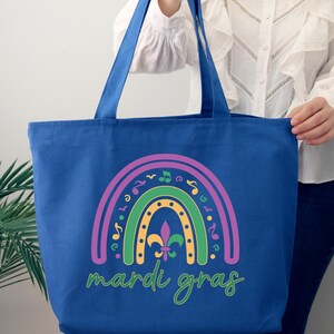 K&ouml;nnte beinhalten: Eine blaue Canvas-Tragetasche mit einem Regenbogen-Design mit Musiknoten und einer Fleur-de-lis. Der Text "Mardi Gras" ist unter dem Regenbogen gedruckt.