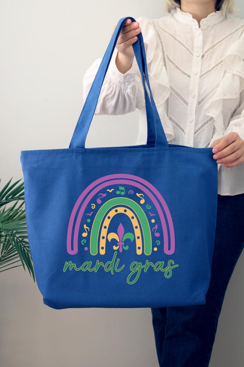 K&ouml;nnte beinhalten: Eine blaue Canvas-Tragetasche mit einem Regenbogen-Design mit Musiknoten und einer Fleur-de-lis. Der Text "Mardi Gras" ist unter dem Regenbogen gedruckt.
