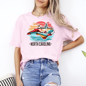 May include: A pink t-shirt with a graphic of the state of North Carolina in a vintage style with a sunset and cityscape. The text "North Carolina" is printed below the graphic.