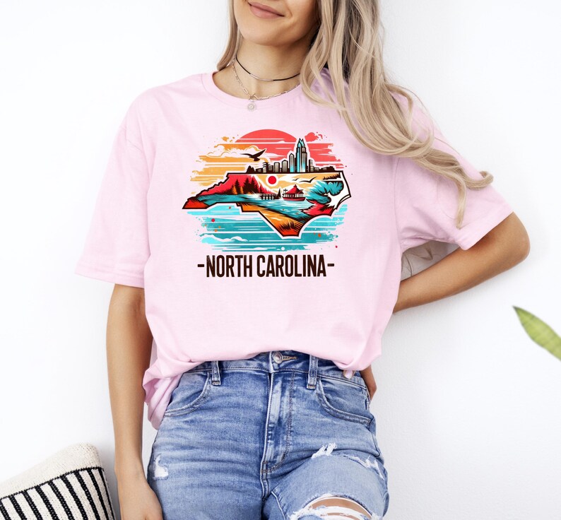 May include: A pink t-shirt with a graphic of the state of North Carolina in a vintage style with a sunset and cityscape. The text "North Carolina" is printed below the graphic.