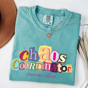 May include: A turquoise t-shirt with the words "Chaos Coordinator" in a colorful, cut-out style design. The phrase "classroom edition" is printed below. A gold necklace with a pendant rests on the shirt.