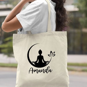 May include: A natural canvas tote bag with a black silhouette of a person meditating in a crescent moon with a lotus flower. The bag has the name "Amanda" printed in black below the image.