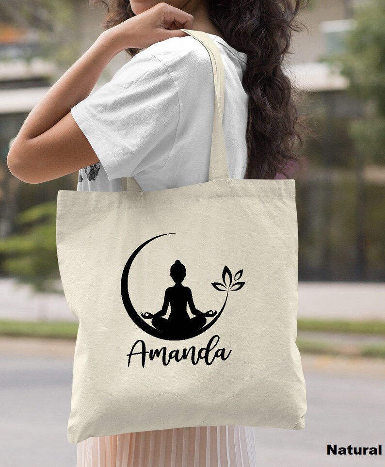 May include: A natural canvas tote bag with a black silhouette of a person meditating in a crescent moon with a lotus flower. The bag has the name "Amanda" printed in black below the image.