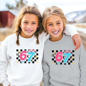 May include: Two children wearing sweatshirts. One is white, the other is gray. Both sweatshirts have a graphic design with the number "67" in pink and blue, the word "SEVEN" and a black and white checkered background.