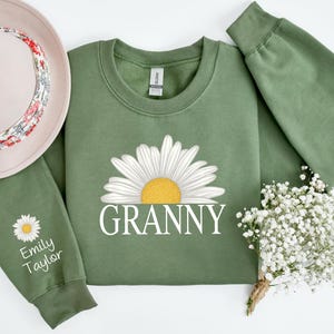 May include: Green sweatshirt with a white daisy and the word "Granny" printed on the front. The sweatshirt has a white daisy on the sleeve with the name "Emily Taylor" printed below it.