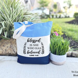 May include: A tri-color blue tote bag with white canvas and a black quote that reads "blessed is she who has believed" with the scripture reference "LUKE 1:45".