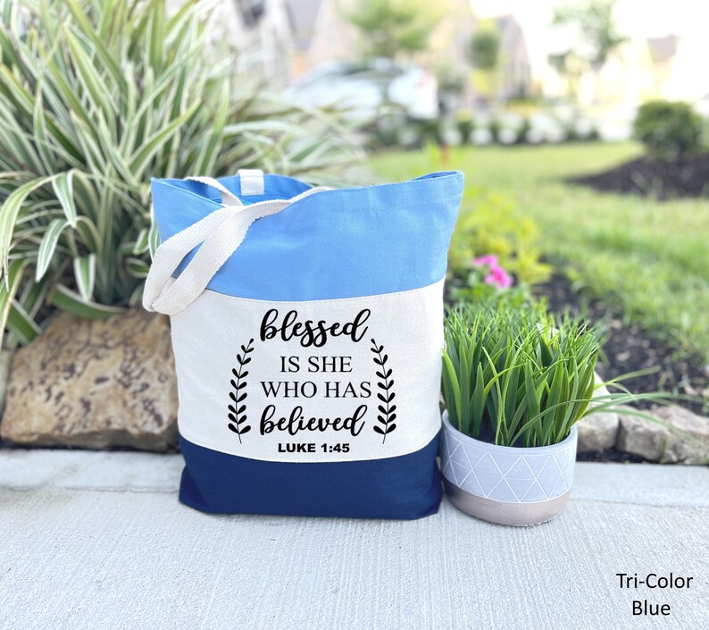 May include: A tri-color blue tote bag with white canvas and a black quote that reads "blessed is she who has believed" with the scripture reference "LUKE 1:45".