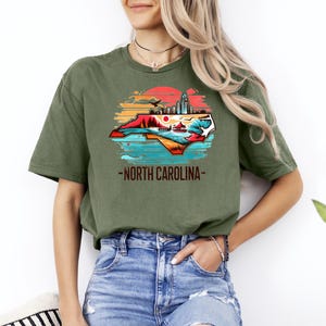 May include: A green t-shirt with a graphic of the state of North Carolina. The graphic features a sunset, a cityscape, and a beach scene. The text "North Carolina" is printed below the graphic.