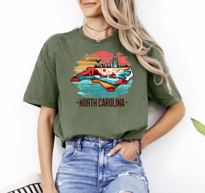 May include: A green t-shirt with a graphic of the state of North Carolina. The graphic features a sunset, a cityscape, and a beach scene. The text "North Carolina" is printed below the graphic.