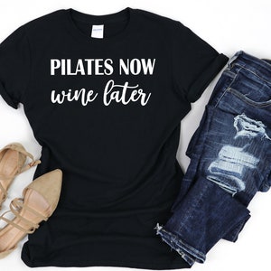 K&ouml;nnte beinhalten: Schwarzes T-Shirt mit wei&szlig;em Text, der "Pilates Now Wine Later" lautet. Das Shirt wird mit einer blauen Jeans und braunen Sandalen kombiniert.