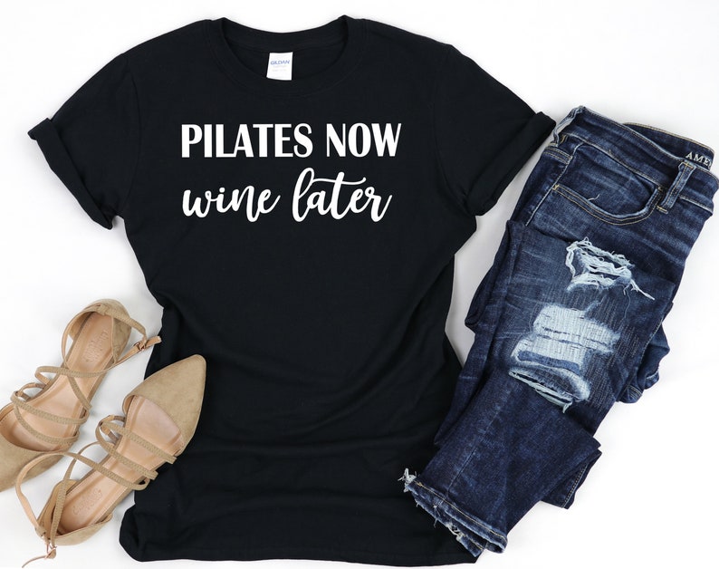 K&ouml;nnte beinhalten: Schwarzes T-Shirt mit wei&szlig;em Text, der "Pilates Now Wine Later" lautet. Das Shirt wird mit einer blauen Jeans und braunen Sandalen kombiniert.